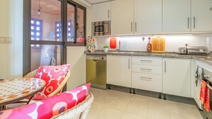 Uma cozinha compacta com armários brancos, micro-ondas embutido e máquina de lavar louça. Há um espaço de refeições com mesa de vime e almofadas coloridas.