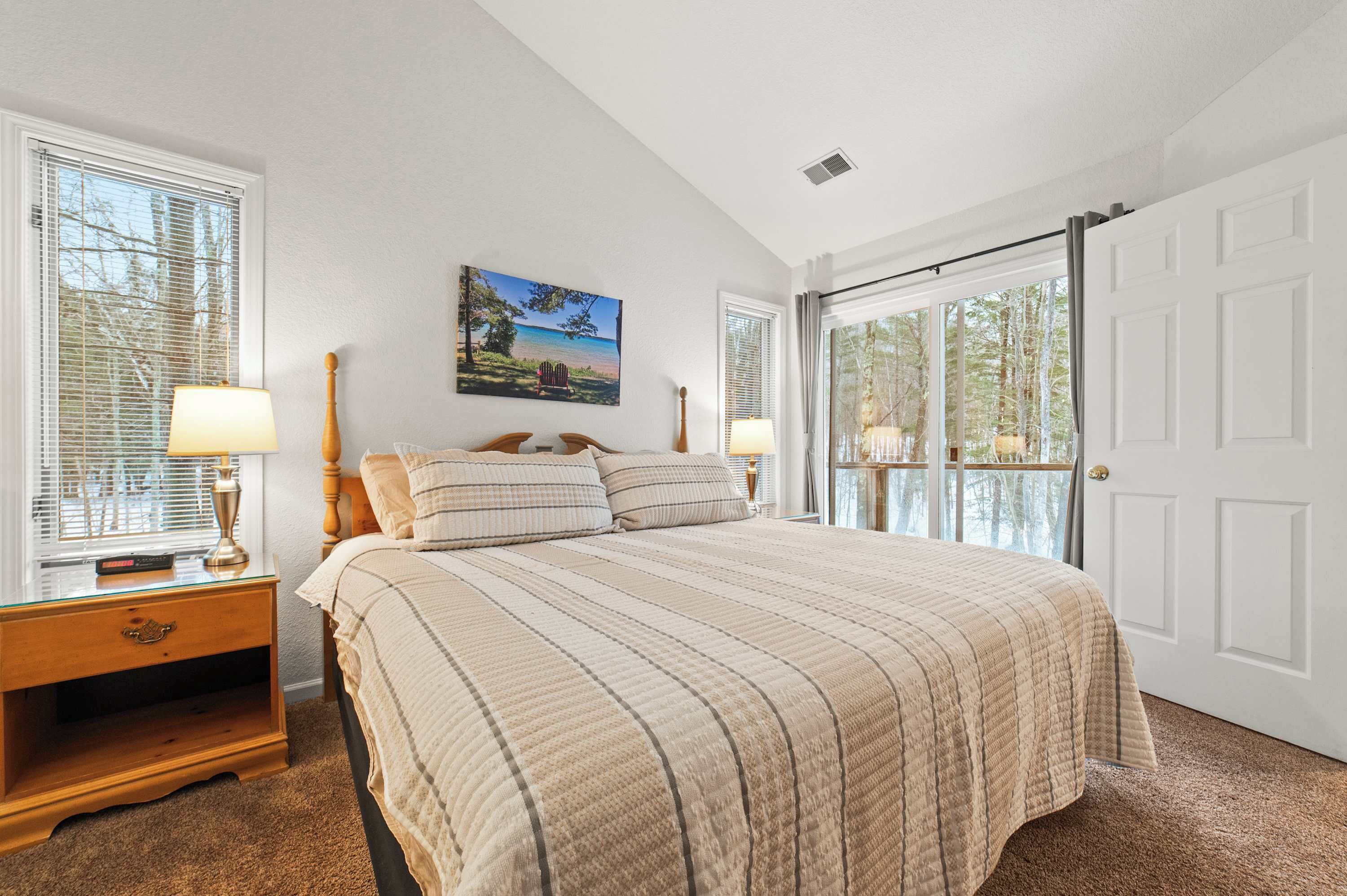 A bedroom with a large bed, two bedside tables with lamps, a painting on the wall, and a sliding glass door leading to a balcony.