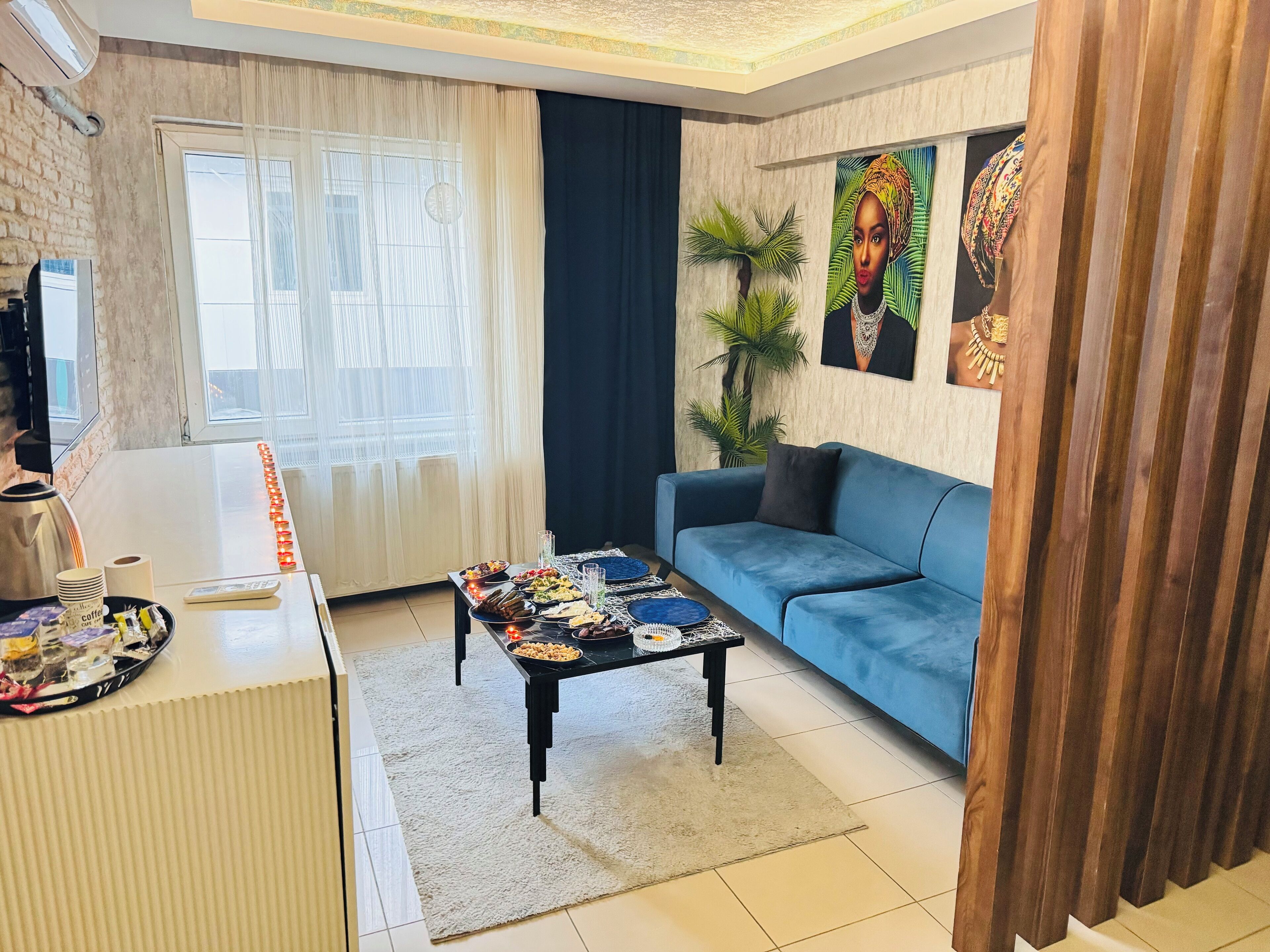 Una sala de estar moderna con un sofá azul, una mesita de centro con aperitivos, un televisor y un ventanal con cortinas ligeras.