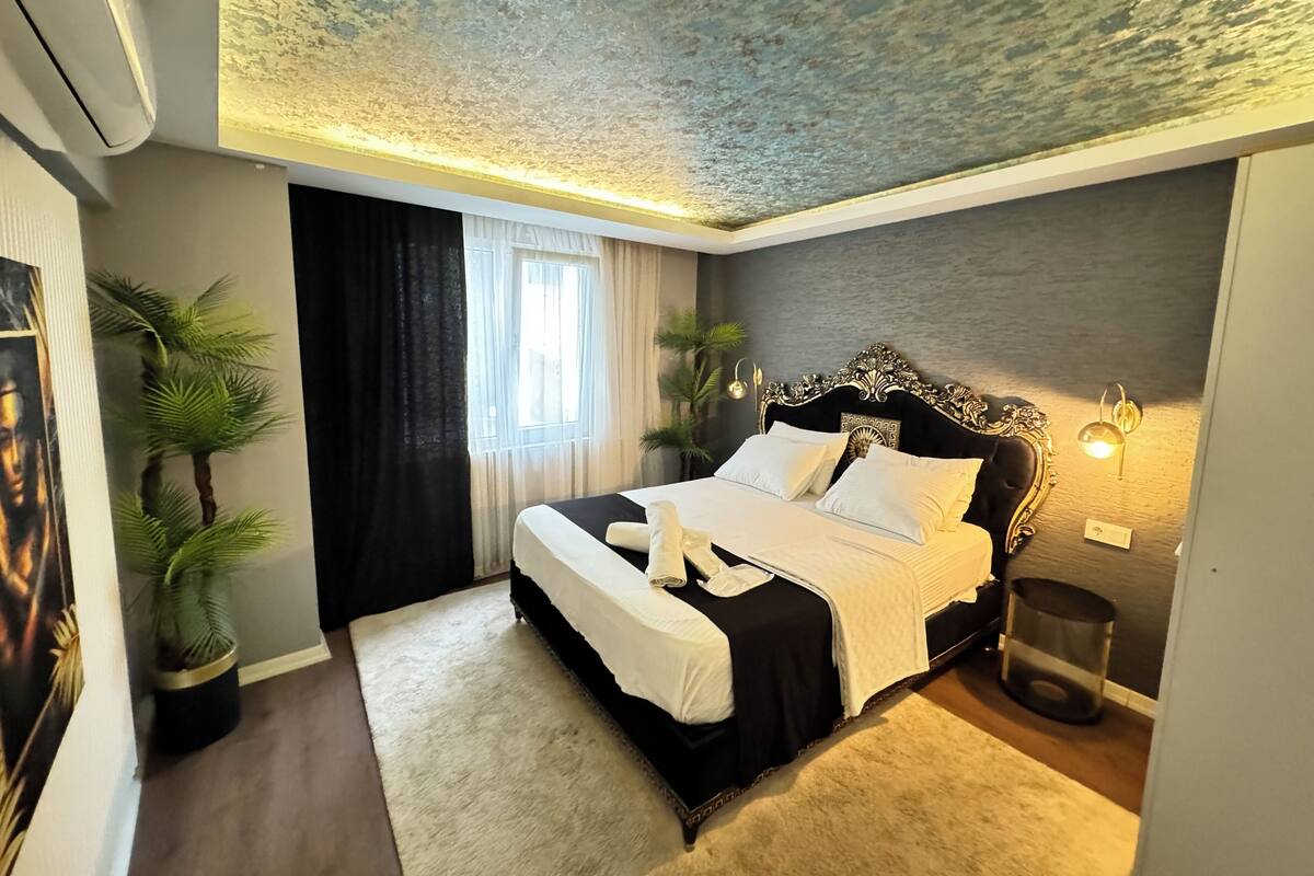 A modern bedroom with a large bed, a decorative headboard, a ceiling with a textured finish, and a window with sheer curtains.