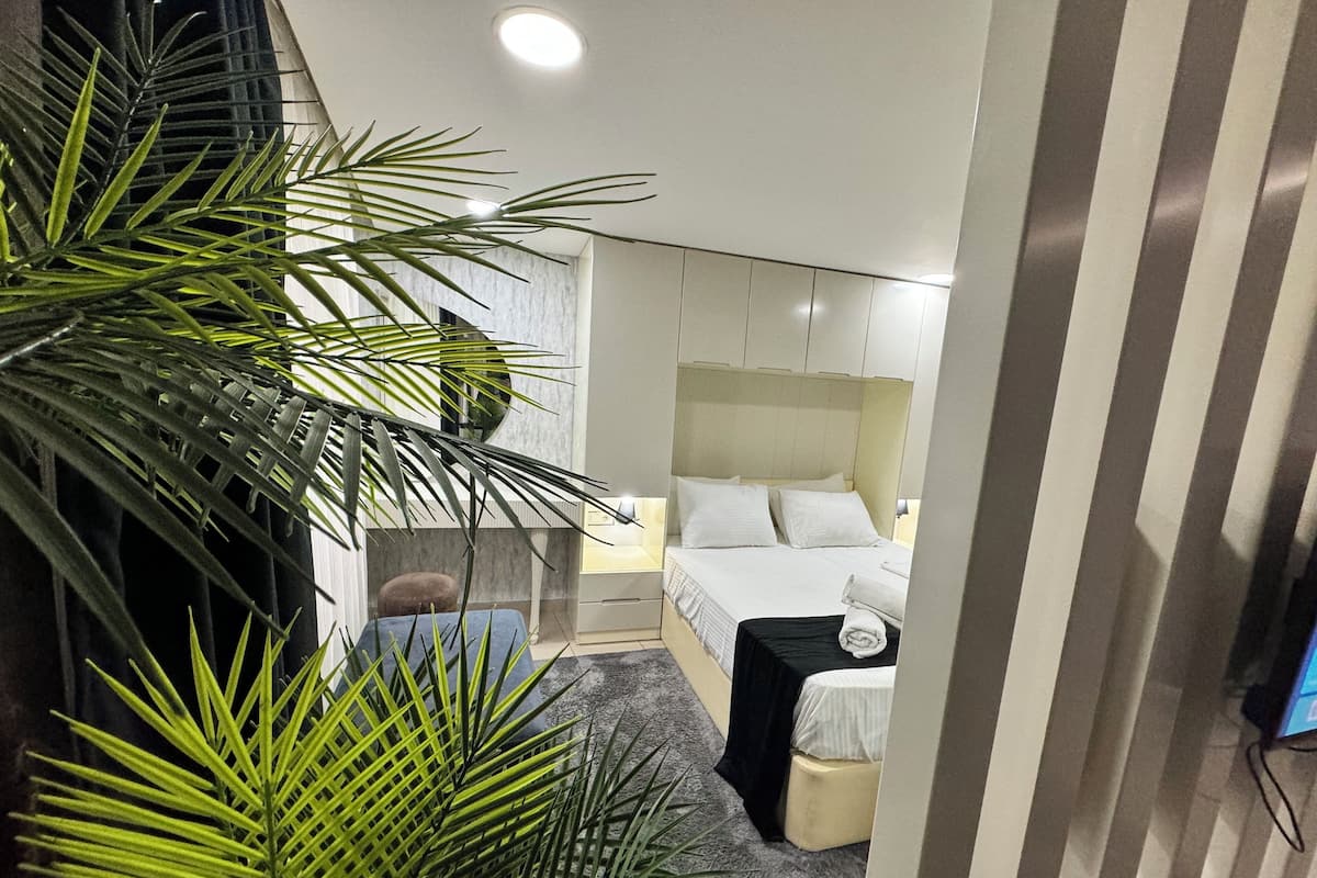 A modern hotel room with a large bed, a small sofa, a nightstand, and a potted plant.