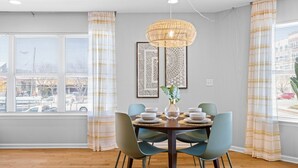 Un coin repas avec une table ronde dressée pour quatre personnes, entourée de chaises de couleur turquoise, d'un luminaire suspendu en rotin et de deux œuvres d'art encadrées accrochées au mur.
