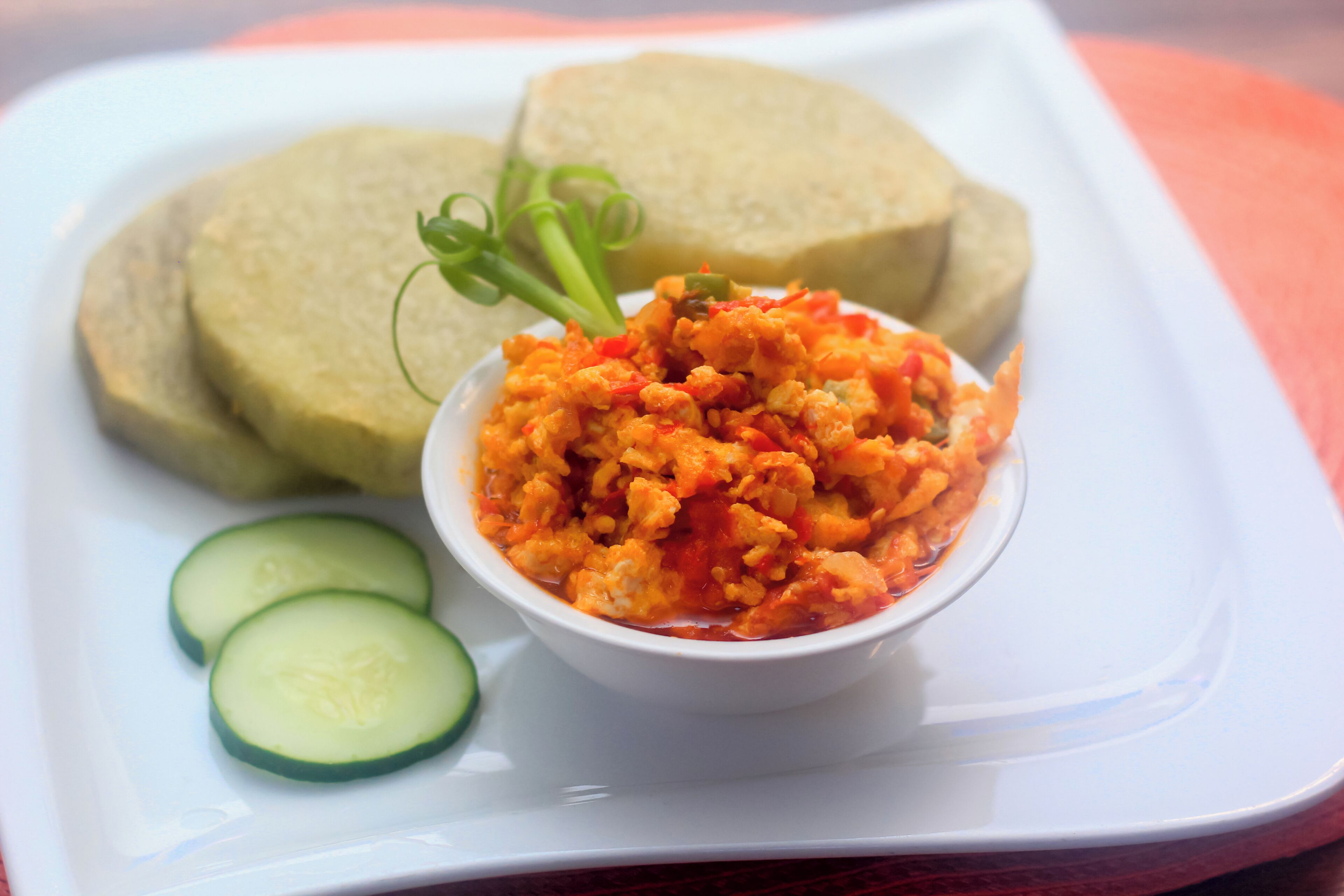 A white plate with a bowl of orange-colored food, slices of cucumber, and two pieces of bread.