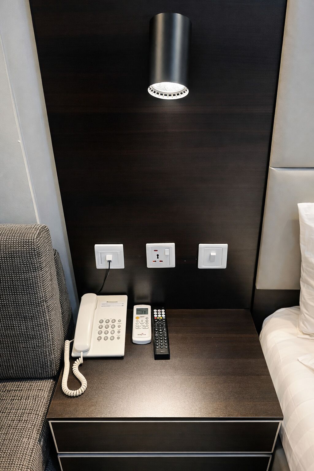 A hotel room with a dark wood headboard, a bedside lamp, a phone, a remote control, and a power outlet.