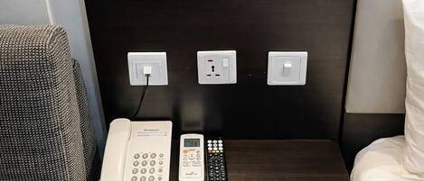 Habitación de hotel con cabecera de madera oscura, lámpara de noche, teléfono, control remoto y toma de corriente.