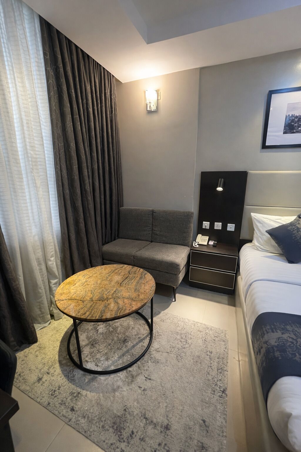 A hotel room with a bed, a sofa, a small round table, a nightstand, a wall-mounted phone, and a framed picture on the wall.