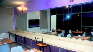 A bar area with a long counter, multiple chairs, and tables. There is a TV mounted on the wall and a window providing a view outside. - Five Clover Inn Abijo (Lekki)