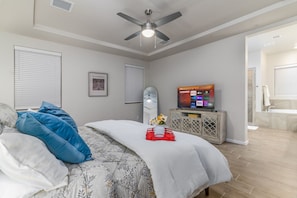A bedroom with a bed, a ceiling fan, a TV, and a bathroom visible through an open door.