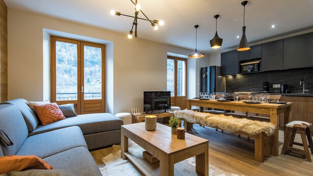 A modern living room with a sofa, a wooden coffee table, and a dining area with a bench. There is a kitchen with dark cabinets and a large window. - ☆☆☆ 3 Vallées & cycling cols – Cosy 6 to 8 stay (Orelle)