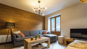 A modern living room with a wooden wall, a grey sofa, a coffee table, a TV, and a large window. - ☆☆☆ 3 Vallées & cycling cols – Cosy 6 to 8 stay (Orelle)