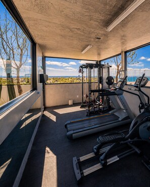 A gym with treadmills, a weight bench, and a squat rack, with a view of palm trees and a body of water through large windows.
