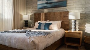 Cama bem arrumada com manta estampada, duas mesas de cabeceira com abajures, cabeceira texturizada e um quadro de um veleiro na parede.