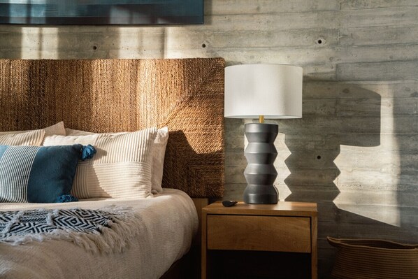 A bedroom with a bed, pillows, a bedside table, a lamp, and a woven wall headboard.