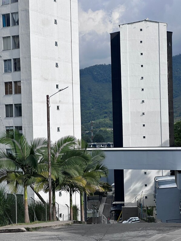 A tall, modern building with multiple windows and a flat roof, surrounded by palm trees and a hilly landscape in the background.