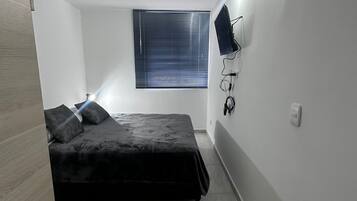 A bedroom with a bed, a window with blinds, a wall-mounted TV, and a light fixture.