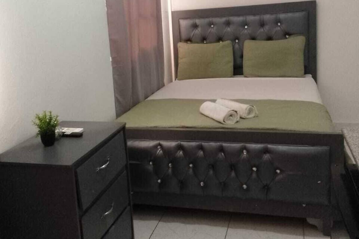 A bedroom with a dark upholstered bed, a bedside table with drawers, a small plant, and a curtain.