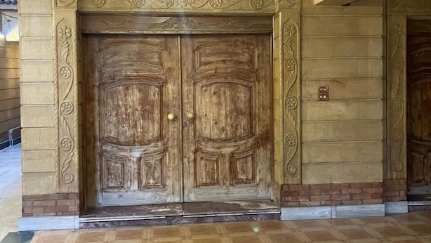 Una gran puerta doble de madera con intrincados grabados, un accesorio de iluminación arriba y un piso a cuadros.