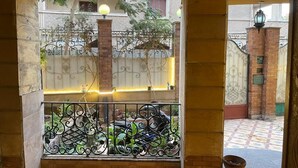 Un balcón con barandillas metálicas ornamentadas, una planta en maceta y vista a un muro de ladrillo y una ventana.