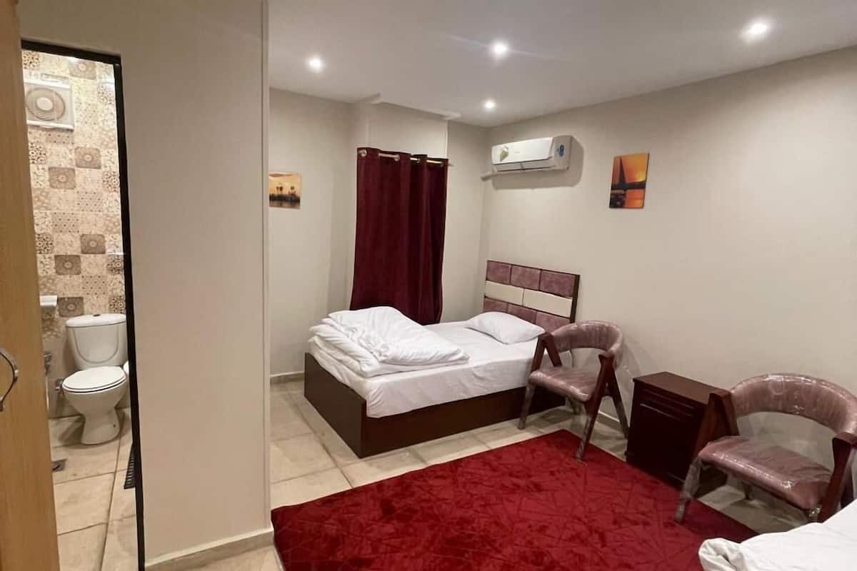 A bedroom with a bed, a chair, a red rug, an air conditioning unit, and a toilet visible through an open door.