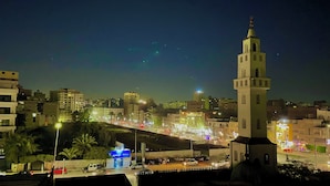 Un paisaje urbano nocturno con una imponente torre de mezquita, una calle concurrida con vehículos y edificios iluminados.