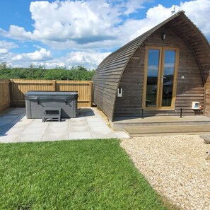 A wooden cabin with a sloped roof, a hot tub, and a patio area with a wooden deck and stone tiles. - Cosy Rural Cabin with Electric Hot Tub (Grantham)