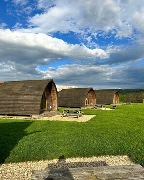 A row of rustic wooden cabins with pitched roofs, arranged in a grassy field with a clear sky above. - Cosy Rural Cabin with Electric Hot Tub (Grantham)
