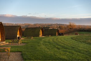 A row of wooden cabins with picnic tables in a grassy field, set against a backdrop of trees and a cloudy sky. - Cosy Rural Cabin with Electric Hot Tub (Grantham)