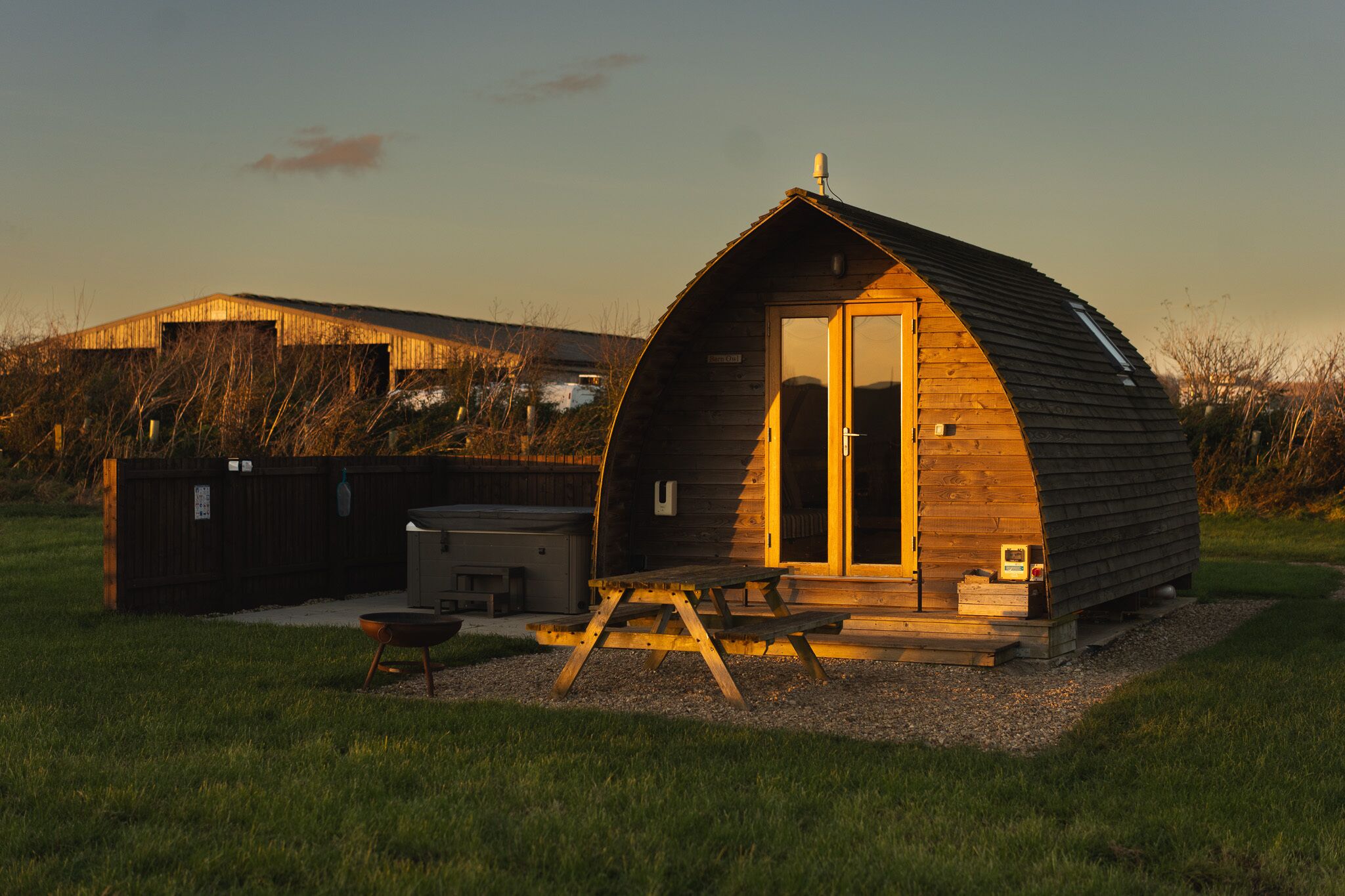 Een klein, houten vakantiehuisje met een gebogen dak, een deur en een klein terras met een tafel en stoelen.