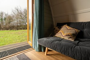A room with a dark grey sofa, a patterned throw pillow, a window with a view of a grassy area, and a curtain. - Cosy Rural Cabin with Electric Hot Tub (Grantham)