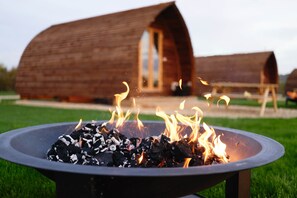 A fire pit with burning wood in the foreground, surrounded by a grassy area. In the background, there are wooden structures with arched roofs. - Cosy Rural Cabin with Electric Hot Tub (Grantham)