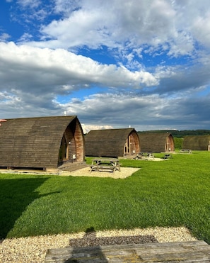 A row of rustic wooden cabins with pitched roofs, arranged in a grassy field with a clear sky above.