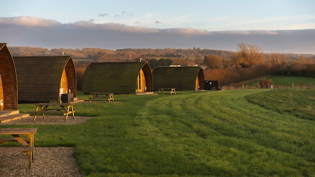 A row of wooden cabins with picnic tables in a grassy field, set against a backdrop of trees and a cloudy sky.