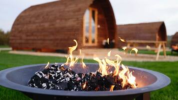 A fire pit with burning wood in the foreground, surrounded by a grassy area. In the background, there are wooden structures with arched roofs.