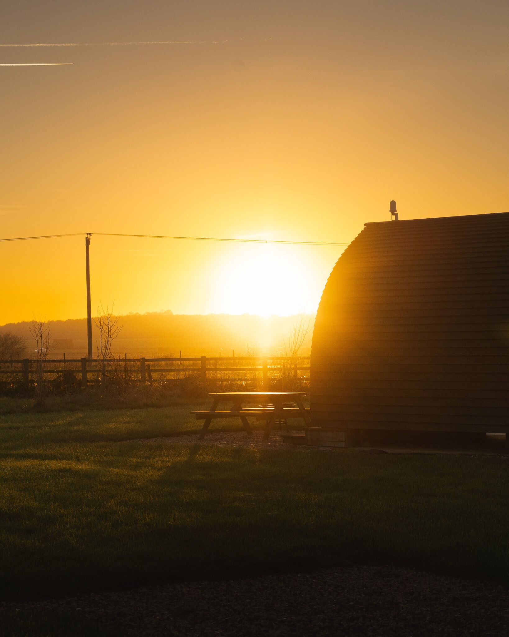 Ein Sonnenuntergang über einer ländlichen Landschaft mit einem Bauernhaus, einem Holzzaun und einem Picknicktisch.