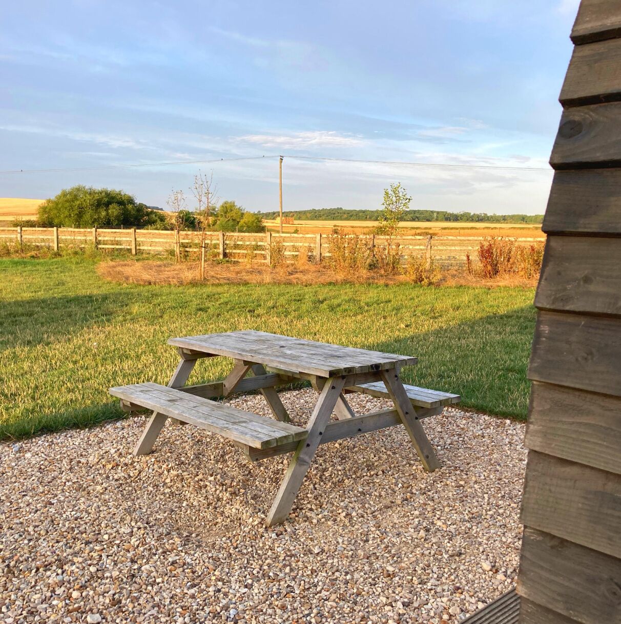 Una mesa de picnic de madera en un área de grava, con un paisaje rural de fondo.