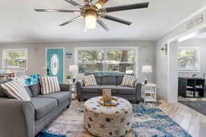 A living room with two grey sofas, a round ottoman, a ceiling fan, and a door with a blue glass panel.