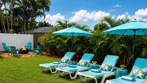 Een achtertuin met een rij witte lounge stoelen en turquoise parasols, omgeven door weelderige begroeiing en een vuurkorf.