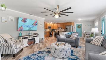 A modern living room with a flat-screen TV, a ceiling fan, and a dining area with a kitchen in the background.