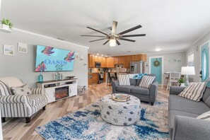 A modern living room with a flat-screen TV, a ceiling fan, and a dining area with a kitchen in the background.