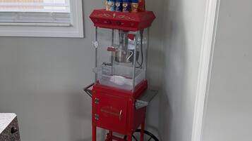 A red popcorn machine with a clear container, placed in a corner with a window and a wall decoration.