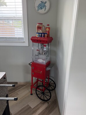 A red popcorn machine with a clear container, placed in a corner with a window and a wall decoration.