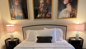 Una cama bien tendida con ropa de cama blanca y una manta azul oscuro, flanqueada por dos mesitas de noche con lámparas, y tres retratos enmarcados sobre la cama.