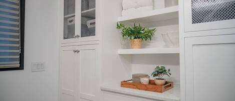 A white bathroom with a window, a towel rack, a shelf with toiletries, and a plant.