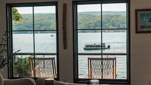 Una habitación con un ventanal grande con vistas a un lago, una silla de madera, una mesa con un libro y un vaso, y un cuadro en la pared.
