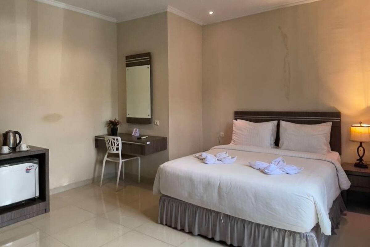 A hotel room with a bed, a desk, a chair, a mirror, and a nightstand with a lamp.