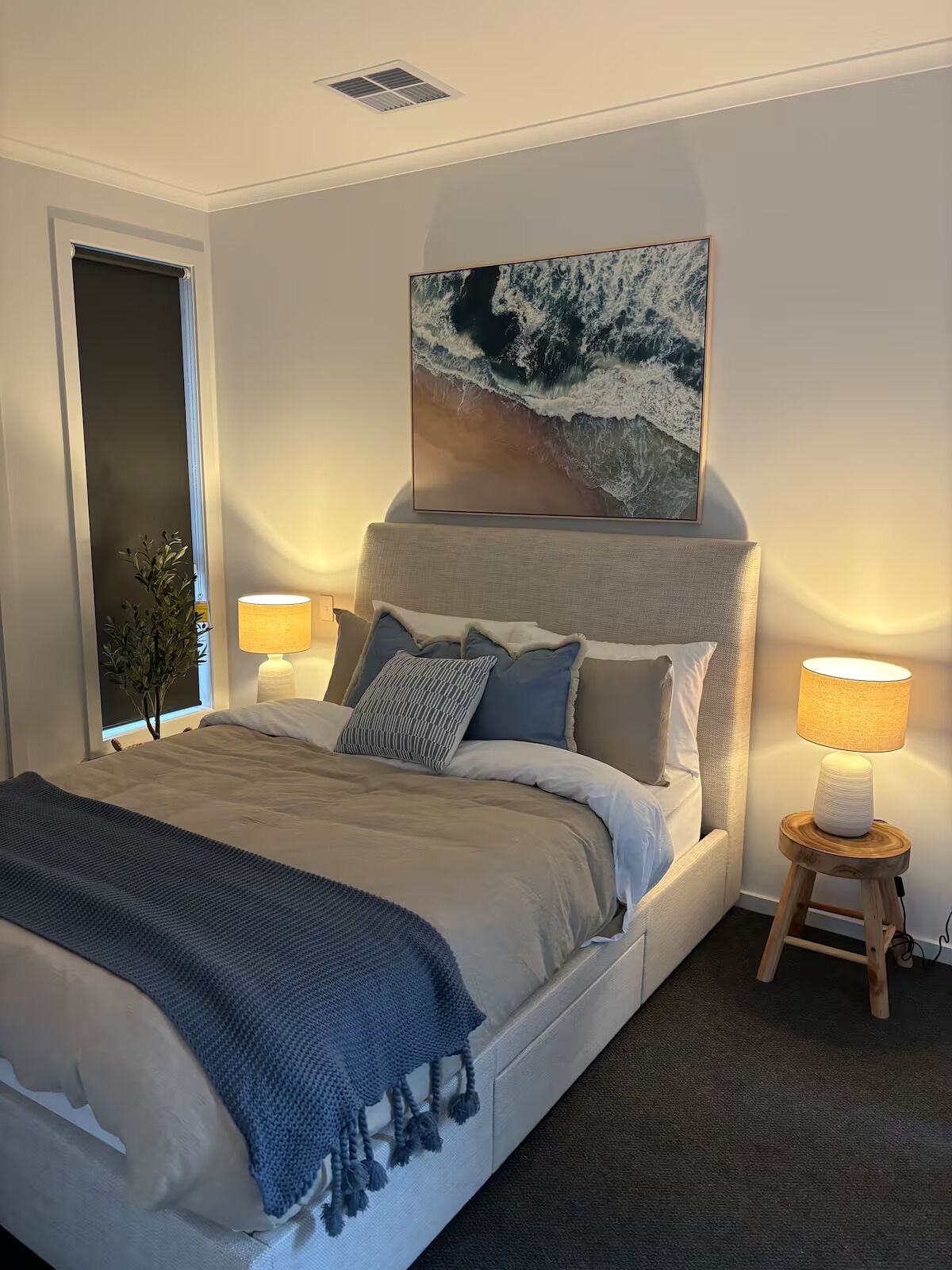 A modern bedroom with a large bed, a headboard with a painting, bedside lamps, and a small wooden side table.
