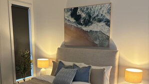 A modern bedroom with a large bed, a headboard with a painting, bedside lamps, and a small wooden side table.
