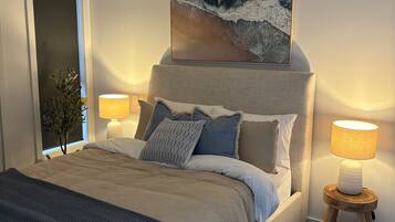A modern bedroom with a large bed, a headboard with a painting, bedside lamps, and a small wooden side table.