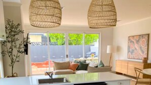 A modern kitchen with a central island, white cabinetry, and a black dishwasher. There are two hanging wicker pendant lights, a dining area with a table and chairs, and a view of a backyard with a fence and greenery.