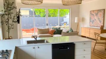 A modern kitchen with a central island, white cabinetry, and a black dishwasher. There are two hanging wicker pendant lights, a dining area with a table and chairs, and a view of a backyard with a fence and greenery.